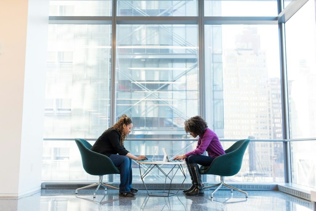 two women against a glass wall in an office environment working on laptop computers