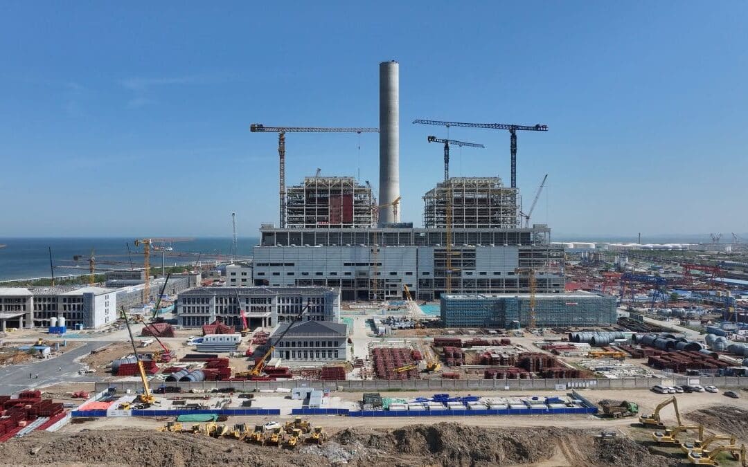 Large-scale thermal power plant under construction with cranes, steel framework, and coastal background
