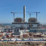 Large-scale thermal power plant under construction with cranes, steel framework, and coastal background