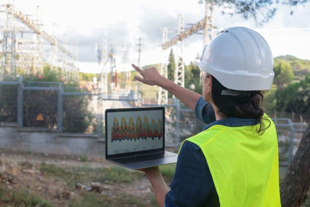 Female electrical engineer in hard hat and safety vest outdoors, holding a laptop with graphs while pointing at a power substation, overseeing grid operations and data analysis